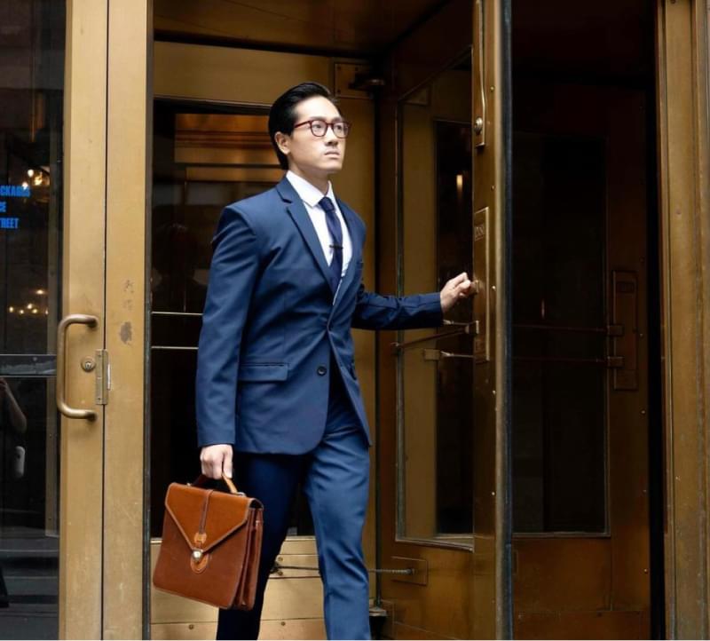 Businessman exiting office building with leather briefcase, wearing navy suit and glasses