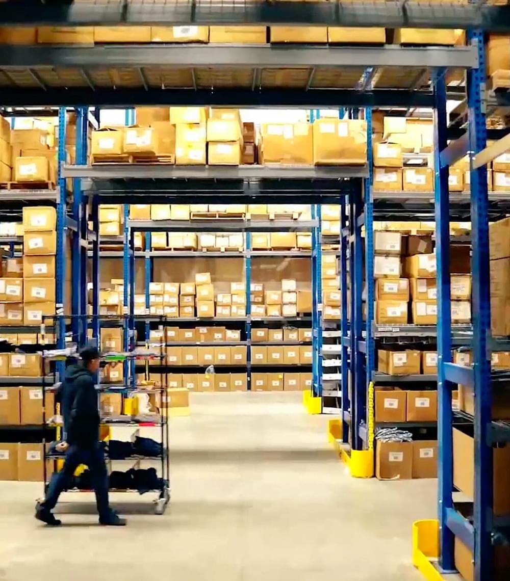 Warehouse worker pushing cart through shelves stocked with cardboard boxes for eCommerce order fulfillment