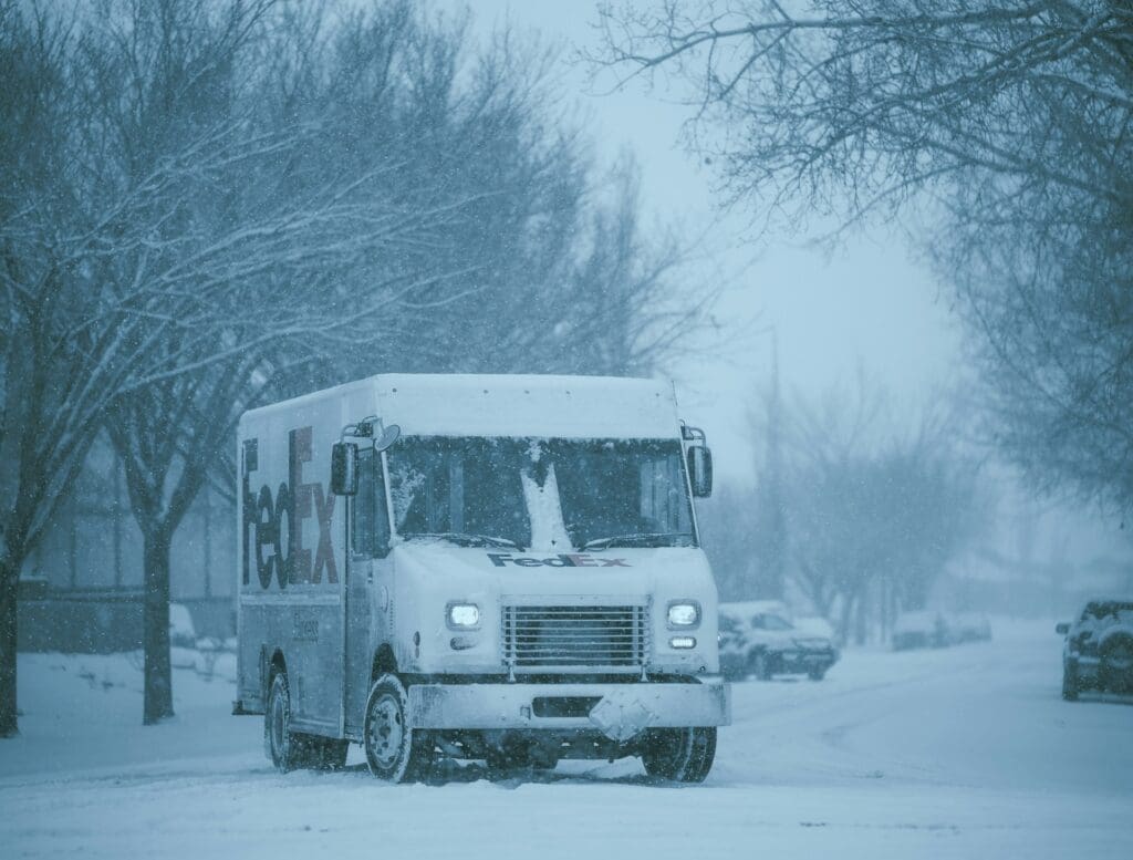 FedEx delivery truck driving through heavy snow on residential street lined with bare trees