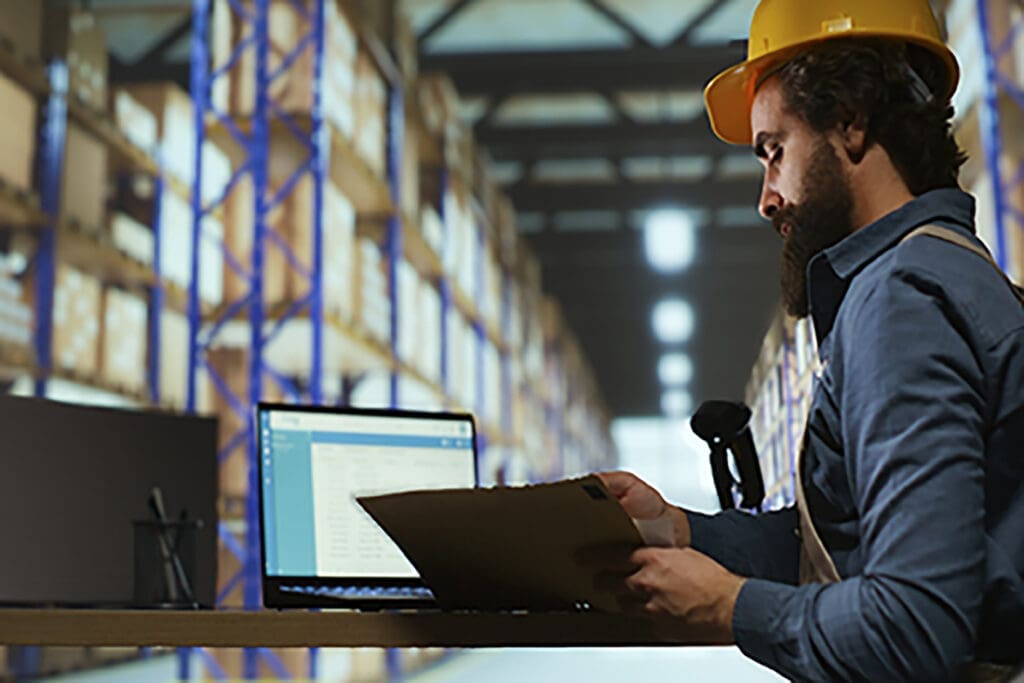 Warehouse worker wearing hard hat using laptop in fulfillment center with high shelves in background