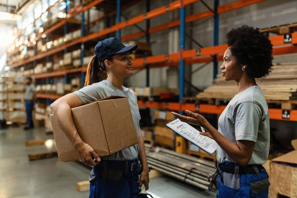 Two female warehouse workers discussing clipboard while holding boxes in a large warehouse.