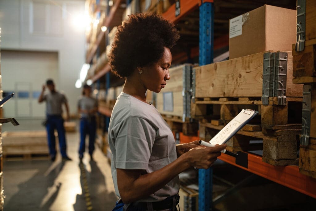 Woman reviewing inventory on clipboard in warehouse with shelves of boxes