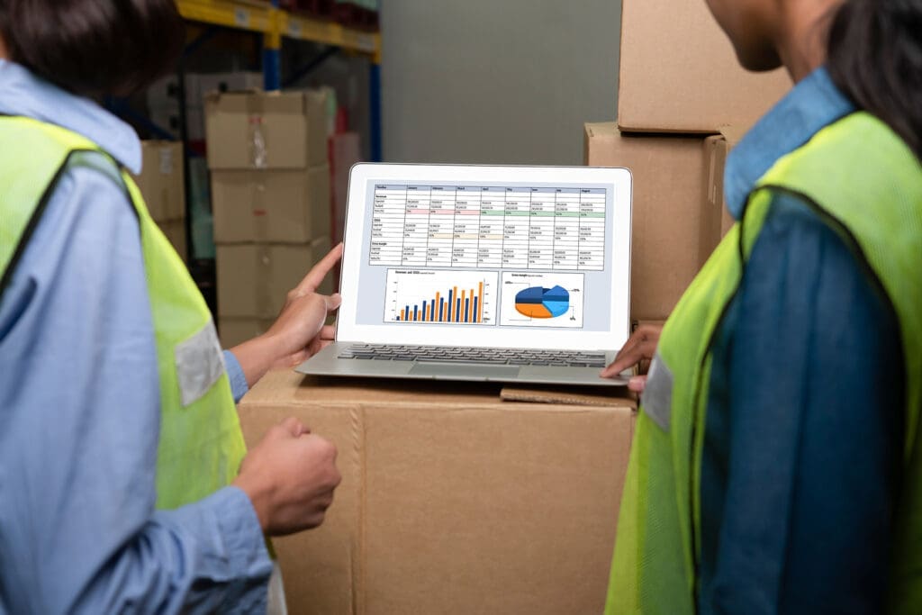 Workers in reflective vests reviewing logistics data on laptop in warehouse with boxes