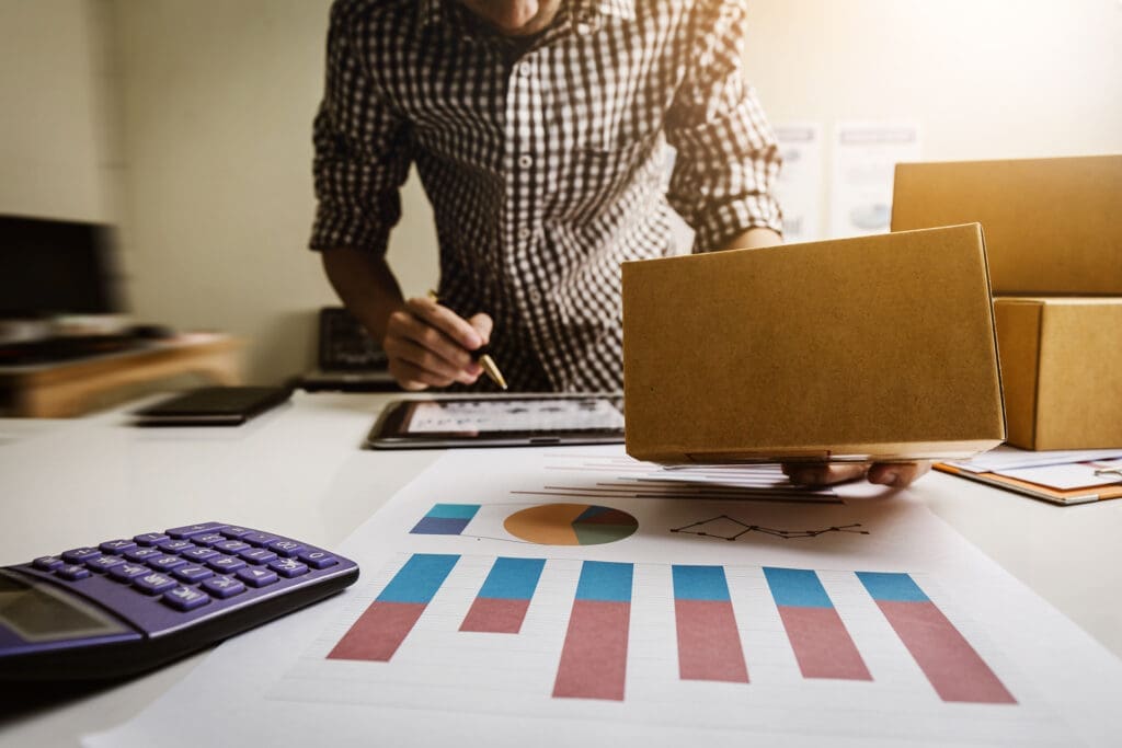 Woman analyzing shipping reports and data on desk with calculator, tablet, and boxes